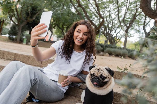Carefree young and curly woman in casual clothes having video call on cellphone and holding coffee to go while sitting near pug dog on stairs in blurred park at daytime in Barcelona, Spain  - Foto, Imagem