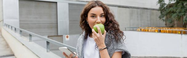 若いですブルネット女性とともに暖かいセーター肩の上に熟した緑のリンゴを食べ、スマートフォンを保持しながら、バルセロナ、スペイン、バナー、工業ビルの街のカメラを見て - 写真・画像
