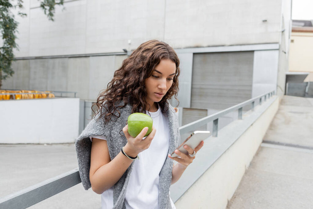Mladá a hezká žena s teplým svetrem na ramenou pomocí mobilu a drží čerstvé jablko, zatímco stojí na rozmazané městské ulici s průmyslovou budovou v Barceloně, Španělsko  - Fotografie, Obrázek