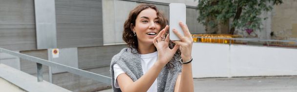 Smiling young curly woman with warm sweater on shoulders having video chat on smartphone while standing on blurred urban street with industrial building at background in Barcelona, Spain, banner  - Foto, immagini