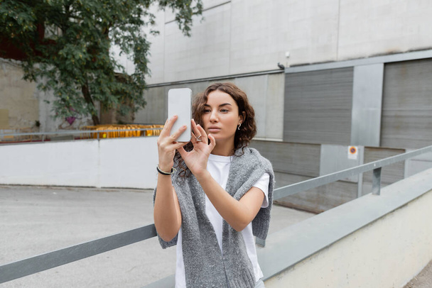 Красуня з теплим светром на плечах за допомогою мобільного телефону стоїть на вулиці з розмитим індустріальним будинком на задньому плані в Барселоні (Іспанія).  - Фото, зображення