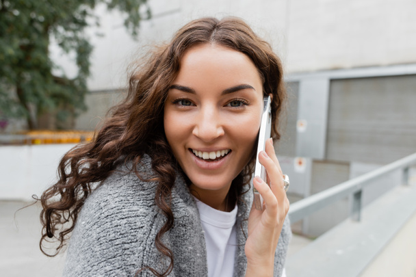 Portrait of positive and young curly woman with sweater on shoulders looking at camera while talking on smartphone on blurred urban street in Barcelona, Spain  - Foto, afbeelding
