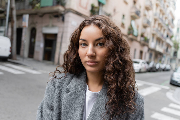 Portrait of young and curly brunette woman in casual grey jacket standing on blurred urban street with buildings at background at daytime in Barcelona, Spain  - Photo, Image