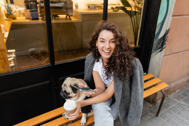 Positive junge brünette Frau in lässiger Jacke mit Smartphone und Kaffee to go, während sie neben dem Mops Hund auf einer Bank in der Nähe eines Straßencafés in Barcelona sitzt, Spanien  - Foto, Bild
