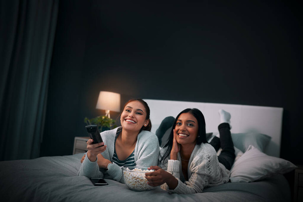 Women friends on bed at night for movies, eating popcorn and happy sleepover at home with streaming service or television. Gen z people relax in bedroom with TV film, cinema subscription and internet. - Fotografie, Obrázek