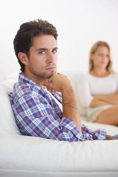 Filled with frustration - relationship issues. A upset man sitting in a sofa with hand on chin and upset wife sitting in the background - Photo, Image