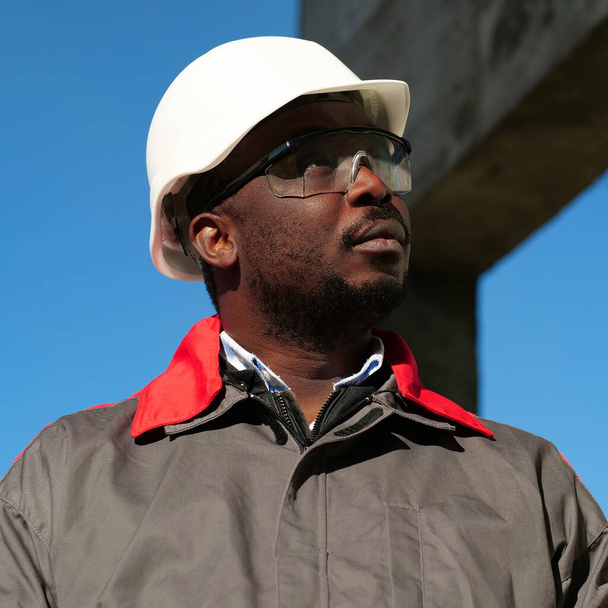 Afrykański amerykański pracownik w twardej czapce i okularach ochronnych stoi na placu budowy - Zdjęcie, obraz