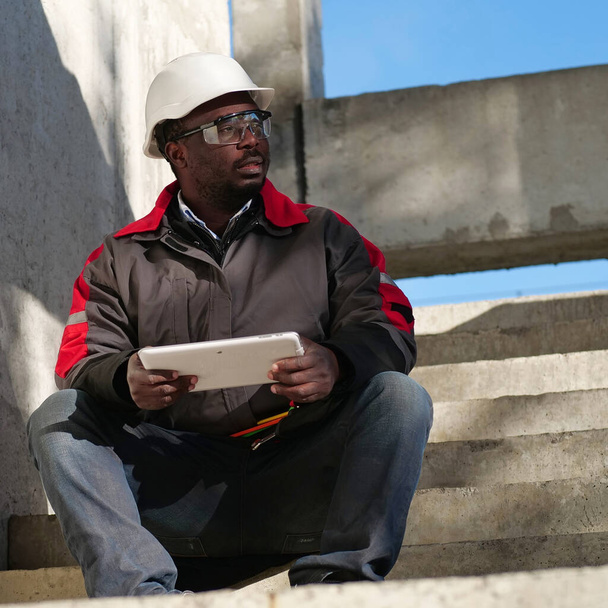 Moe Afro-Amerikaanse werknemer met half gesloten ogen in witte harde hoed zit op de bouwplaats en houdt in handen tablet PC - Foto, afbeelding