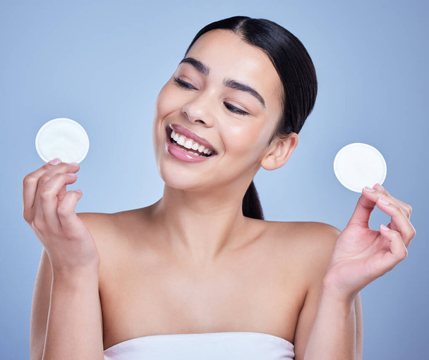A beautiful smiling mixed race woman using a cotton pad to remove makeup during a selfcare grooming routine. Hispanic woman applying cleanser to her face against blue copyspace background. - Foto, imagen