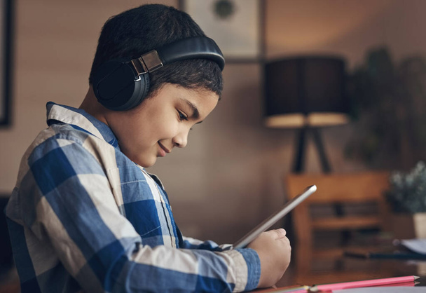 Powiązany z szerszą społecznością wychowawczą. uroczy chłopiec korzystający z tabletu cyfrowego i słuchawek podczas wykonywania zadania szkolnego w domu - Zdjęcie, obraz