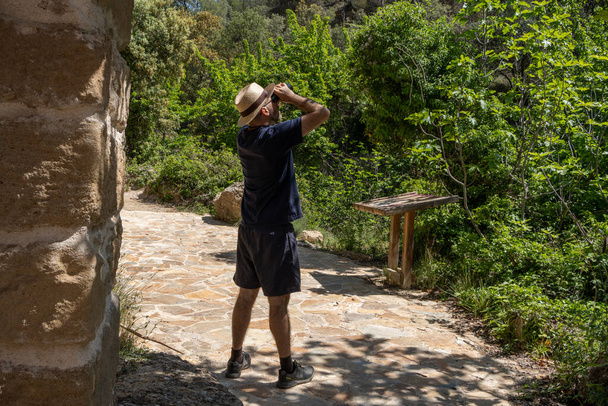 man in an explorer's cap looking through binoculars in a natural area, in the middle of a rural wash in a forest, ornithological tourist looking at the birds in the trees. - Photo, Image
