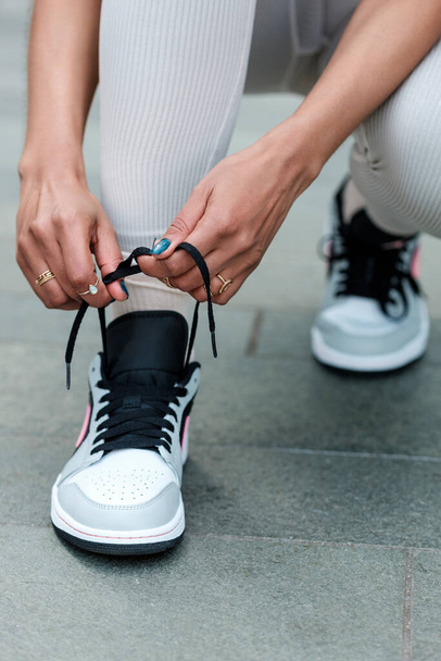 Unrecognizable fitness woman tying her shoelaces outdoors on grey rough floor tiles. Blue nails and jewelry on fingers. - 写真・画像