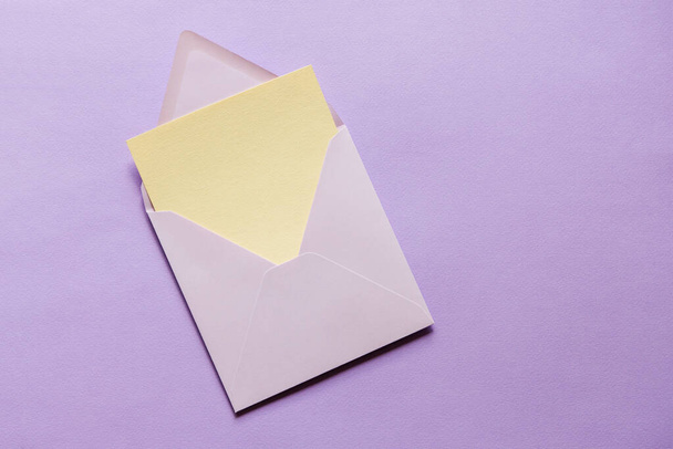 空のグリーティングカードがモックアップ。紫色の紙の背景に紫色の封筒に紙のモックアップのベージュの空のシート。平置きだ。結婚式ビジネス誕生日静物画. - 写真・画像
