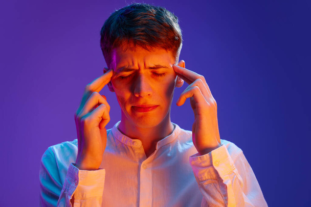 Portrait of young guy in white shirt touching head with hands suffering from headache and overworking against gradient purple background in neon light. Concept of human emotions, lifestyle, youth - Fotoğraf, Görsel