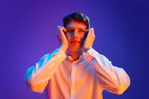 Portret van jonge emotionele man in wit hemd die het gezicht met de handen aanraakt, lijdend aan overbewerkingen en stress tegen gradiënt paarse achtergrond in neon licht. Concept van menselijke emoties, levensstijl - Foto, afbeelding
