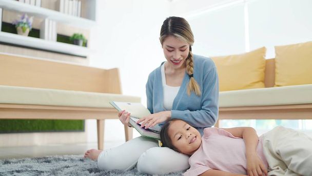 Happy Asian little daughter laying on mother's lap while mother reading fairy tale book sitting floor in living room at home. Family and love concept - Фото, зображення
