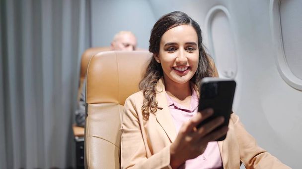 Happy young Hispanic latino woman passenger sitting on seat using smartphone while traveling by airplane. Woman travel on long flight - Photo, Image