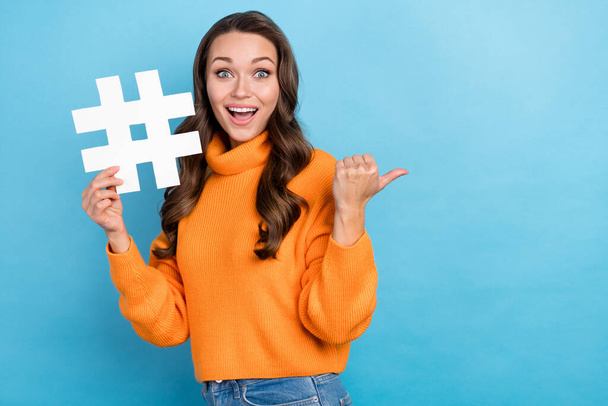 Photo of sweet excited woman dressed orange sweater holding hashtag thumb empty space isolated blue color background. - Foto, Bild