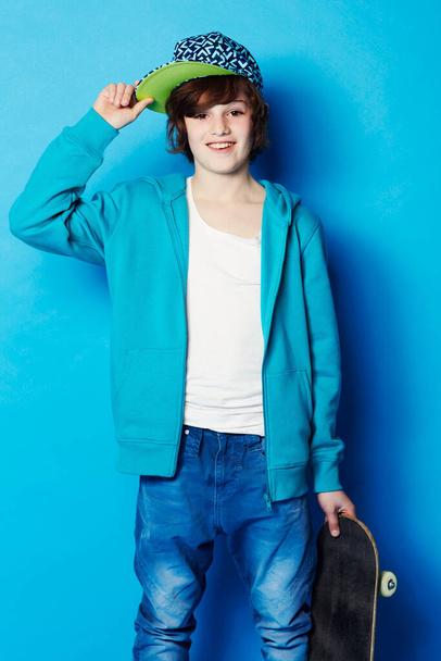 Fashion, skateboard and portrait of a child in a studio with a casual, trendy and cool outfit. Skateboarder, smile and happy boy kid model with skating style posing isolated by a blue background - Foto, Bild