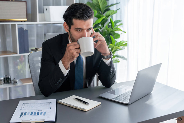 Zakenman werkt in een moderne kantoorruimte met een kop koffie in zijn hand, terwijl hij telefoneert met verkooppraatjes of leidinggevende medewerker. Vurig - Foto, afbeelding