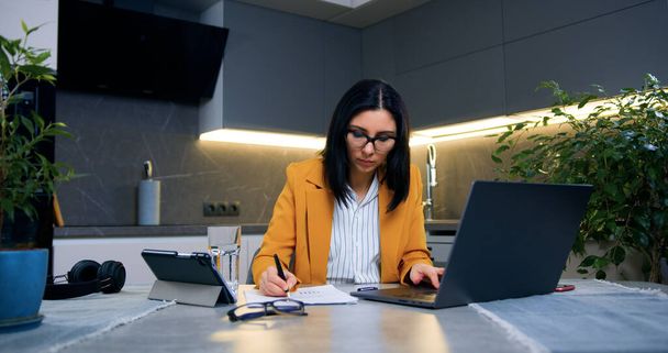 Привлекательная серьезная профессиональная темноволосая женщина в деловой одежде сидит перед компьютером и работает с информацией на экране и сообщает дома - Фото, изображение