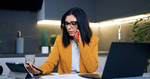 Portrait of good-looking concentrated skilled 35-aged businesswoman in glasses and formal clothes which talking on phone while working with documents and tablet device at home - Foto, Bild