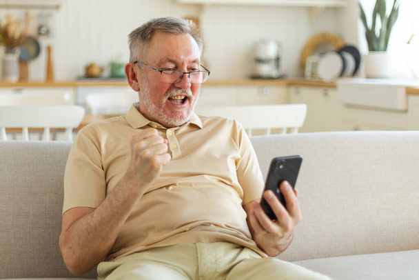 Старшеклассник среднего возраста, обладатель эйфории со смартфоном. Взрослый дедушка смотрит на сотовый телефон чтение отличные новости, получая хороший результат, выиграв онлайн ставку чувство изумления. Жест победы - Фото, изображение