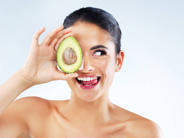 No need to splurge to gain that all over glow. Studio shot of an attractive young woman covering her eye with an avocado against a gray background - Foto, afbeelding