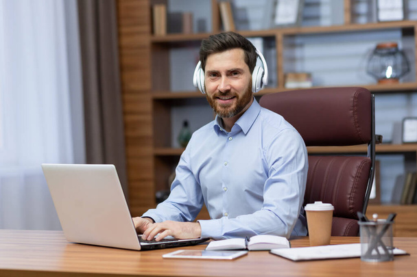 Portrait of a young male businessman working in the office at a laptop, sitting in white headphones and smiling at the camera. - Fotografie, Obrázek