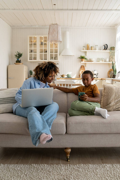 Mãe afro-americana trabalhando no laptop e olhando para o smartphone em filhos mãos sentadas no sofá em casa. Menino adolescente navegando no smartphone, assistindo vídeos. Trabalho ocupado mãe, criança gadget vício.  - Foto, Imagem