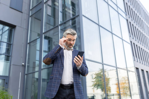 A worried senior man in a suit stands outside an office center and looks disappointedly at the phone screen, putting his glasses down in surprise with his hand on his face. - Photo, image