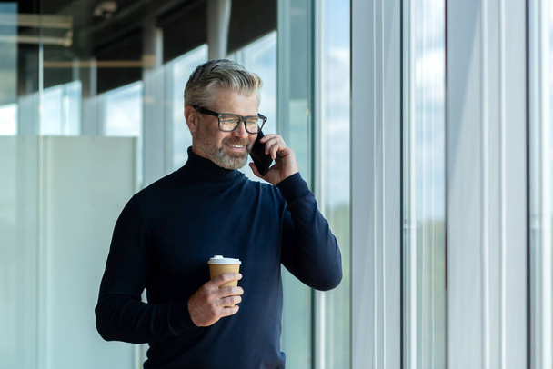 Handsome senior businessman standing in the office center by the window with a cup of coffee with him and smiling talking on the phone. - 写真・画像