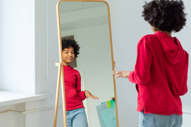 Hou van jezelf. Mooie jonge glimlachende Afrikaanse Amerikaanse vrouw dansend van haar spiegelreflectie. Zwarte dame kijkt naar spiegel die er zelfverzekerd en gelukkig uitziet. Zelfliefde concept - Foto, afbeelding