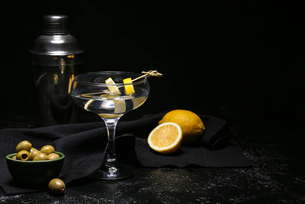 Ένα ποτήρι νόστιμο μαρτίνι, λεμόνια, ελιές και σέικερ σε σκούρο φόντο - Φωτογραφία, εικόνα