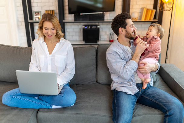 Father carrying infant daughter while mother using laptop on sofa remote work husband helping his wife with baby child. - Foto, imagen