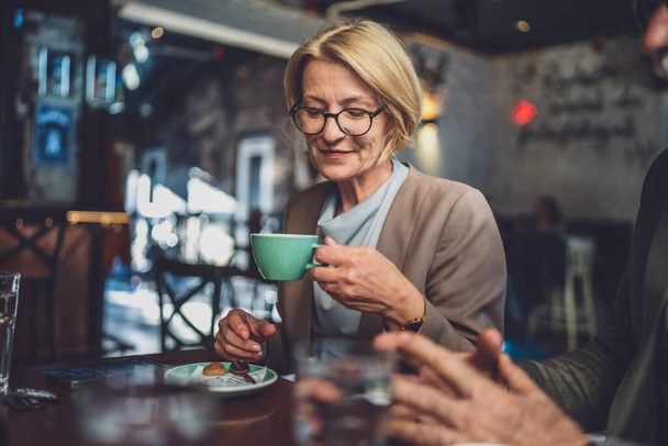 Mature caucasian woman sit in serene ambiance of a cozy dark cafe With a cup of coffee in hand wear eyeglasses enjoy the moment real person copy space - Photo, Image