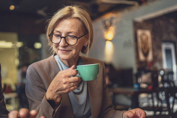 Mature caucasian woman sit in serene ambiance of a cozy dark cafe With a cup of coffee in hand wear eyeglasses enjoy the moment real person copy space - Фото, изображение