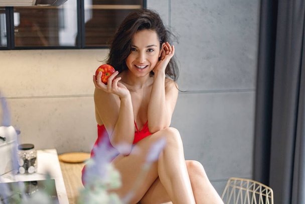 Feliz señora sexy posando mientras está sentado en la mesa en la cocina y sosteniendo la manzana. Mujer bonita vistiendo peignoir rojo - Foto, imagen