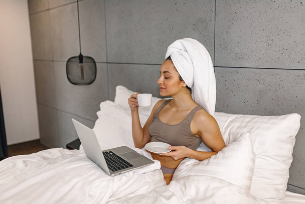 Lachende mooie vrouw met handdoek op haar hoofd zittend in bed terwijl ze koffie drinkt en laptop gebruikt. Communicatie- en technologieconcept - Foto, afbeelding