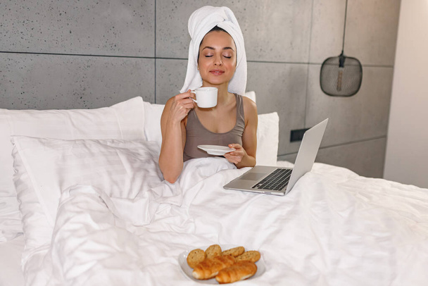 Young lady with closed eyes drinking coffee while sitting on bed and using laptop. Woman with towel in the head. Work at home online - Foto, afbeelding