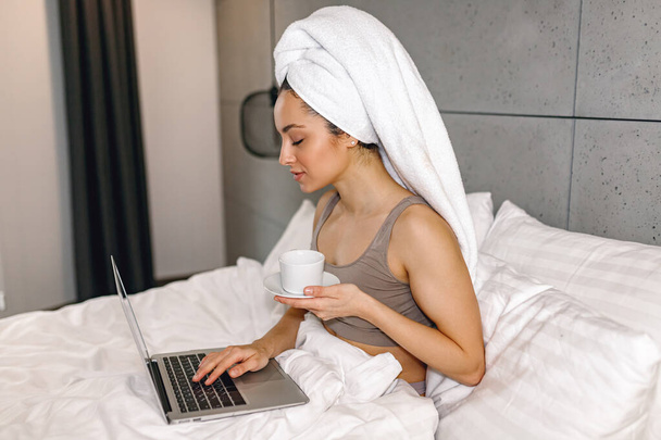 Junge Frau schaut beim Tippen auf Laptop-Bildschirm. Lady genießt morgens eine Tasse Kaffee zu Hause. Lifestyle-Konzept - Foto, Bild