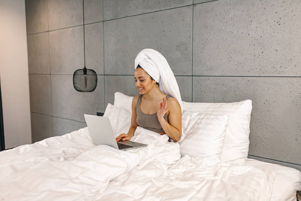 Uśmiechnięta, małostkowa dama machająca ręką podczas rozmowy wideo. Kobieta z ręcznikiem na głowie, używająca laptopa w łóżku. Koncepcja stylu życia - Zdjęcie, obraz