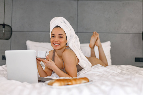 Lachende vrouw genieten van een kopje koffie thuis terwijl ze op het bed in de slaapkamer, met behulp van laptop. Dame met handdoek op het hoofd - Foto, afbeelding