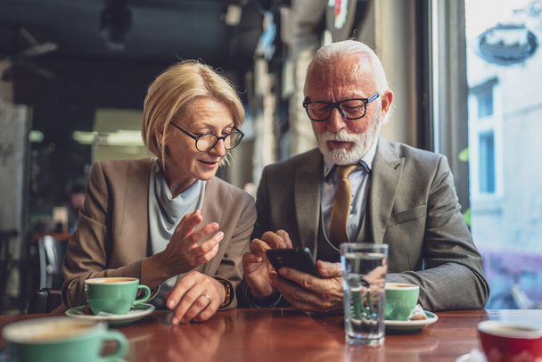 senior man dressed in a suit and a mature blonde woman as business partners husband and wife friends or father daughter talk With smartphones in hand browse internet exchange ideas copy space - Photo, Image