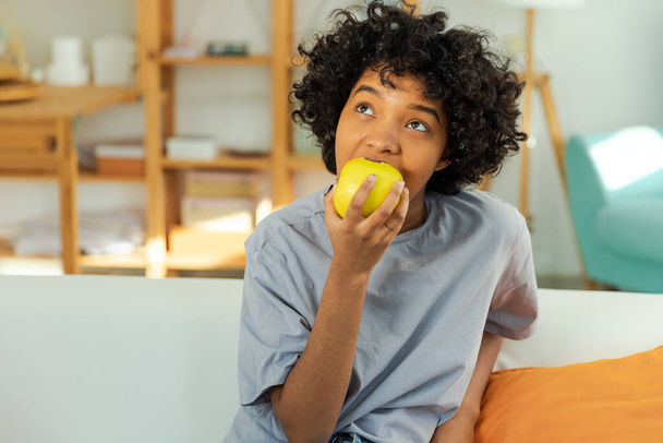 Boldog csinos lány, aki zöld almát harap otthon. Gyönyörű afro-amerikai fiatal nő eszik friss gyümölcsöt és mosolyog. Egészséges étel vegetáriánus diéta koncepció. Egészséges snack tiszta étel - Fotó, kép