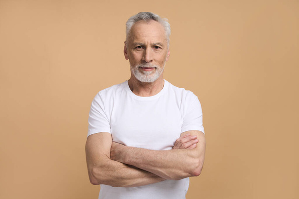 Caucasian confident handsome imposing retired senior man 60 years old, smiling, looking confidently at camera, posing with arms folded isolated on beige background. Mature people and lifestyle concept - Photo, Image