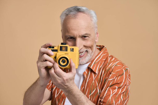 Handsome Caucasian senior man, traveler tourist in orange casual shirt, holding a bright yellow camera, smiling broadly while taking photos, isolated over beige background. People and summer concept - Photo, image