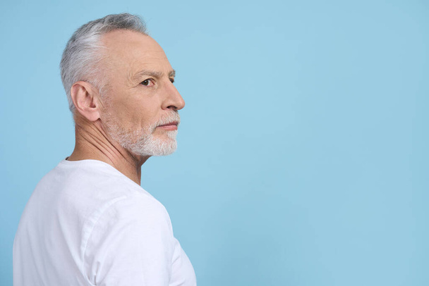 白人の灰色の髪の側の肖像画ハンサムなシニア男性ひげを生やした、退職年金受給者は、慎重に脇を見て、青の背景に隔離された白いカジュアルなTシャツを着ている。広告スペースをコピーします。人々 - 写真・画像
