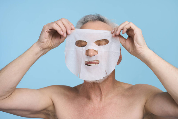 Male beauty and skin care concept. Caucasian senior man putting a hydrating purifying tissue mask on his face, standing with naked torso isolated over blue color background - 写真・画像