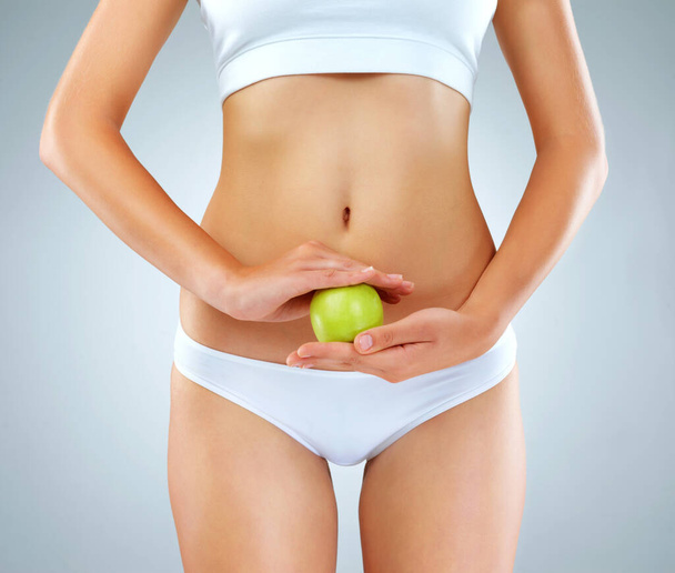 Gesunde Ernährung baut gesunde Körper auf. Eine unkenntliche junge Frau posiert mit einem Apfel vor dem Bauch im Studio - Foto, Bild